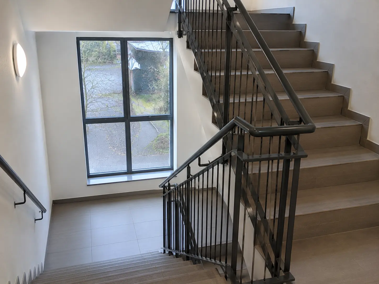 Sauberes Treppenhaus mit dunklem Geländer und großem Fenster