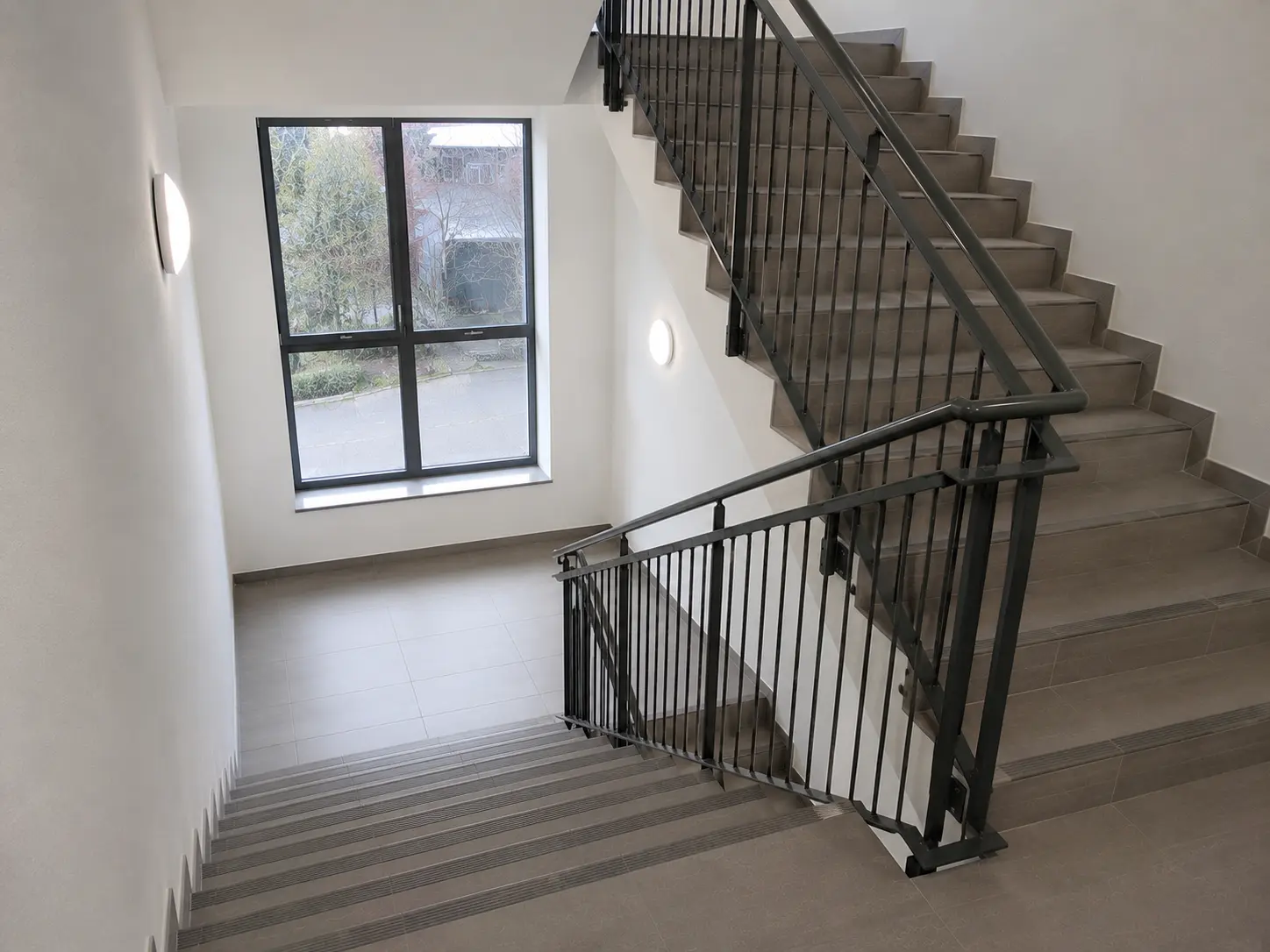Treppenhaus mit gefliesten Stufen, Handlauf an der Wand und Fenster zum Innenhof